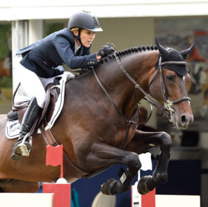 Meredith Michaels en un salto junto a su caballo marrón oscuro. Lleva unos guantes de equitación Roeckl y una americana azul marino.